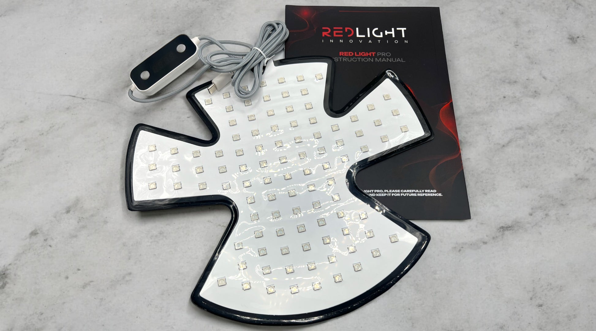 The RedLight Hat Pro LED panel placed on a concrete surface. The panel is rectangular with a grid of red light diodes visible. It is positioned on top of the RedLight Hat Pro instruction manual, which is partially visible underneath. The background is a neutral concrete surface, providing a clear view of both the panel and the manual.