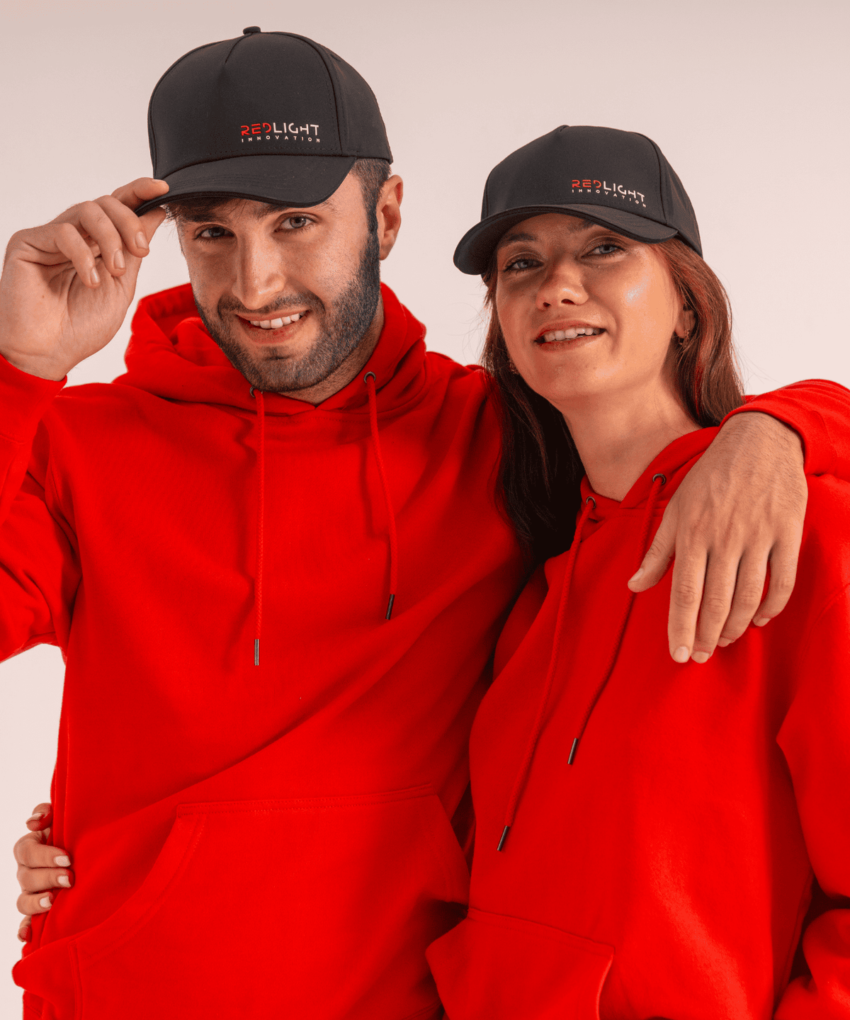 A man and a woman wearing matching bright red hoodies and black red light hats with a small red and white 'REDLIGHT INNOVATION' logo, posed against a plain light background.